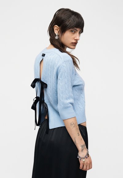 Jeune femme tatouée portant un pull en maille bleu clair avec des rubans noirs noués dans le dos et une jupe noire, regardant par-dessus son épaule.