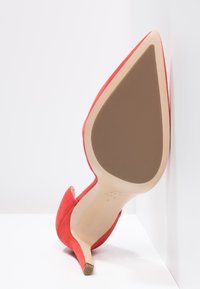 Chaussure à talon haut en daim rouge avec un bout pointu, une silhouette courbée et une semelle beige. Le talon est modérément haut, se rétrécissant vers le bas.