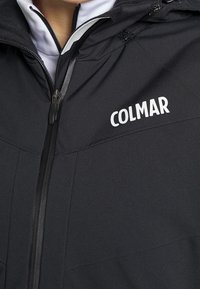 Nahaufnahme einer Person, die eine schwarze Zip-Down-Jacke trägt, auf deren Brust in weißen Buchstaben "COLMAR" gedruckt ist.