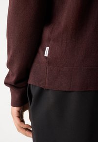 Bordeaux gebreide trui met geribde zoom en manchetten. Heeft een klein merkembleem aan de onderzijde. Gecombineerd met zwarte stoffen broek.
