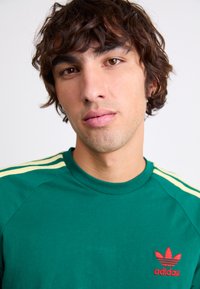 Zeleno bombažno T-shirt z okroundim ovratnikom, rumenimi črtami na ramenih in rdečim logotipom Adidas na levem prsnem delu.