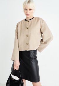 Giacca beige cropped con cinque bottoni dorati e maniche ampie, abbinata a una gonna in pelle nera e a una borsa nera lucida.