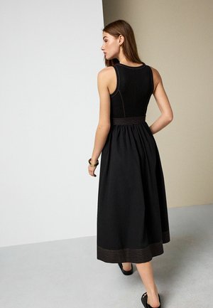 Woman in sleeveless black midi dress with stitched hem and waist detail, standing with one hand on hip, facing away from camera.