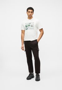 Νέος άνδρας στέκεται φορώντας λευκό T-shirt Lacoste με πράσινο γραφικό κροκόδειλο, μαύρο παντελόνι cargo και μαύρα αθλητικά παπούτσια σε λευκό φόντο.