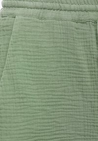 Gros plan d'un tissu vert doux avec une texture matelassée et une taille élastique, montrant une couture latérale et l'ouverture d'une poche.