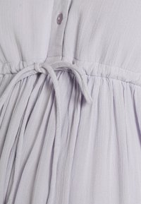 Vestido de lavanda claro com cintura franzida, fecho de botão e tecido texturizado com riscas verticais. As fitas criam uma silhueta ajustada.