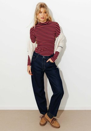 Femme blonde debout, portant un haut à manches longues rayé rouge et noir, un jean bleu foncé, des chaussures marron clair, et un cardigan blanc posé sur les épaules.