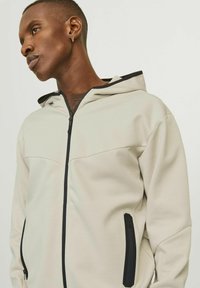Sweat à zip beige clair avec une bordure de capuche noire. Présente une texture lisse et mate ainsi que deux poches zippées à l'avant.