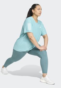 Ljusblå atletisk t-shirt med tre vita ränder på axeln, i kombination med teal leggings och vita träningsskor. Sträckpose.