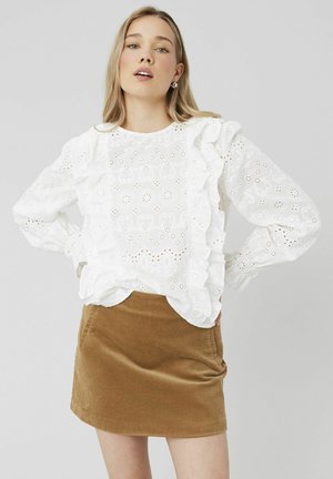 Blouse blanche en coton avec des bords festonnés, dotée de détails en oeillets. Associée à une mini-jupe en velours côtelé marron, au toucher lisse.