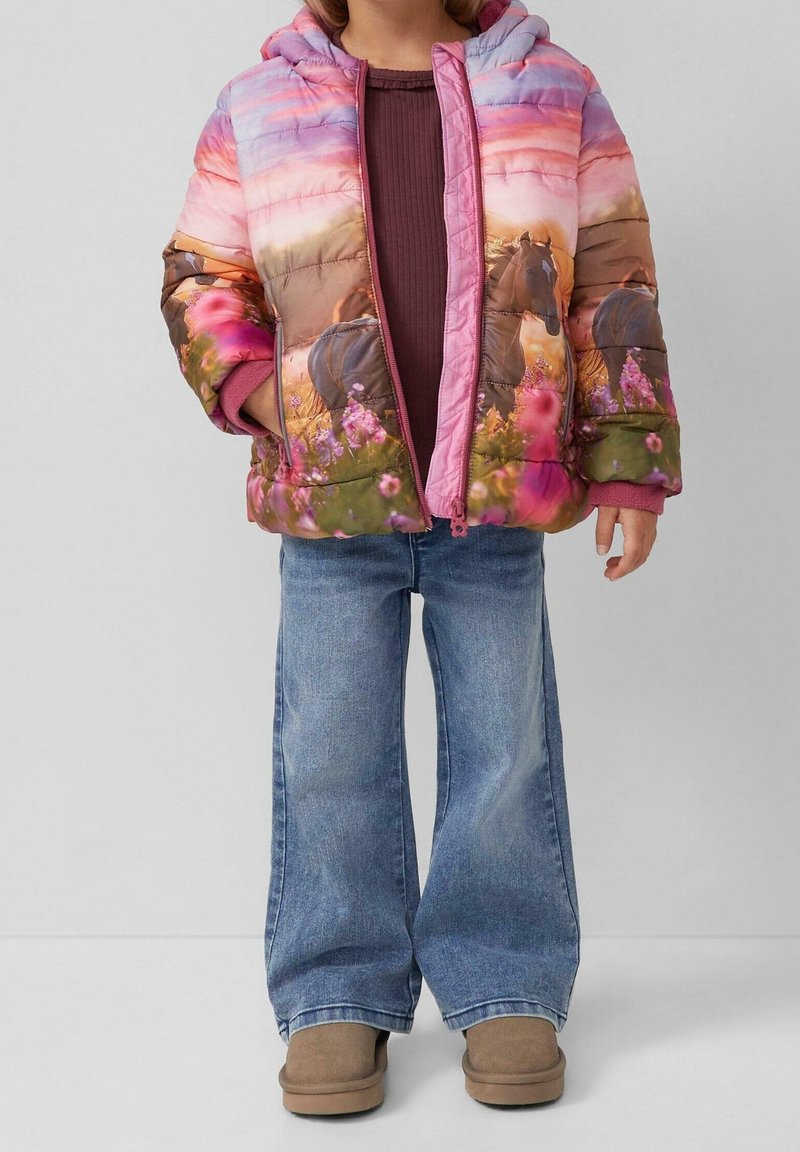 Enfant portant une veste matelassée colorée avec imprimé cheval et fleurs, chemise bordeaux, jean à jambes larges et bottes marron clair, debout devant un fond uni.
