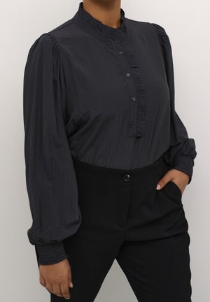 Blouse noire avec patte de boutonnage à volants et poignets froncés, associée à un pantalon noir taille haute, main dans la poche, sur fond uni.