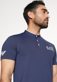 Polo azul marinho com gola abotoada, detalhes brancos e logo EA7 no peito e na manga. Textura suave e ajustada.