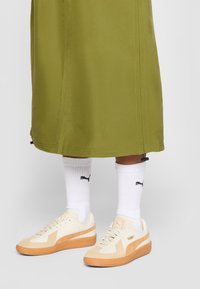 Cream and light beige sneakers with orange rubber soles, paired with white socks featuring a black logo, worn beneath an olive-green skirt.