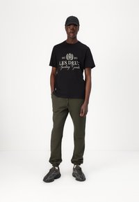 Black cotton t-shirt with "LES DEUX Sporting Goods" graphic, paired with olive green joggers and gray athletic shoes. Black cap included.