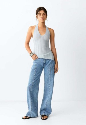 Jeune femme portant un top en maille gris sans manches, un jean bleu à jambes larges, des sandales noires et des bracelets en argent, debout devant un fond blanc uni.