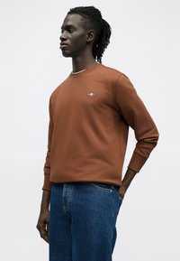 Sweat-shirt marron avec encolure ras du cou, poignets côtelés et logo discret sur la poitrine, associé à un jean en denim bleu. Texture lisse, coupe décontractée.