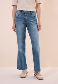 Jeans en denim bleu clair avec une coupe droite, dotés d'une fermeture à deux boutons et de poches avant. Portés avec des baskets blanches et beiges.