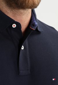 Polo shirt bleu marine en tissu texturé avec un col à motifs. Comprend des boutons blancs et un petit logo rouge, blanc et bleu sur la poitrine.