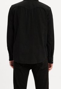 Zwarte spijkerblouse met een puntige schouderpas, lange mouwen en knoopmanchetten, met een gladde textuur en een klassiek kraagontwerp.