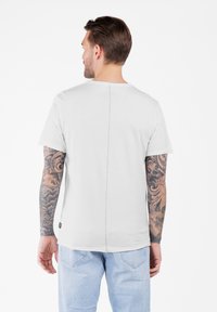 Hombre con los brazos tatuados, vistiendo una camiseta blanca de manga corta y unos jeans azul claro, de espaldas contra un fondo blanco.
