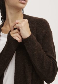 Cárdigan de punto marrón con una textura suave y peluda. Presenta un corte holgado, abertura frontal y puños acanalados, superpuesto a una camisa blanca.