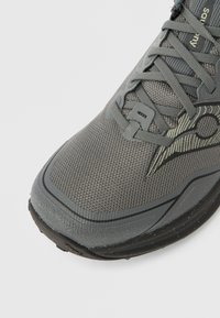 Zapatilla deportiva gris con parte superior de malla, superficie texturizada, puntera redondeada y suela negra contrastante. Cuenta con sistema de cordones y detalle de logotipo.