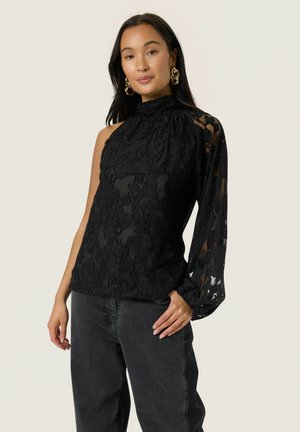 Mujer con blusa negra de encaje de un solo hombro con manga larga y vaqueros gris oscuro, posando frente a un fondo claro y liso.