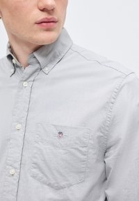 Homme portant une chemise à manches longues gris clair avec une poche poitrine ornée d'un petit logo brodé.