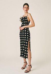 Frau trägt ein schwarzes Midikleid mit großen weißen Punkten, hohem Schlitz am Oberschenkel und dünnen Trägern, kombiniert mit schwarzen Knöchelriemen-Absätzen.