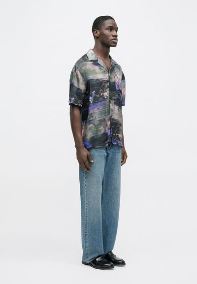 Κοντομάνικο πουκάμισο με πολύχρωμο αφηρημένο σχέδιο, σε άνετη γραμμή. Συνδυάζεται με παντελόνι τζιν με φαρδιά μπατζάκια σε ξεθωριασμένο μπλε χρώμα και μαύρα παπούτσια.