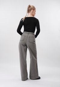 Kvinne med flette, iført svart genser med lange ermer og grå vide bukser med store baklommer, stående mot en ensfarget lys bakgrunn.