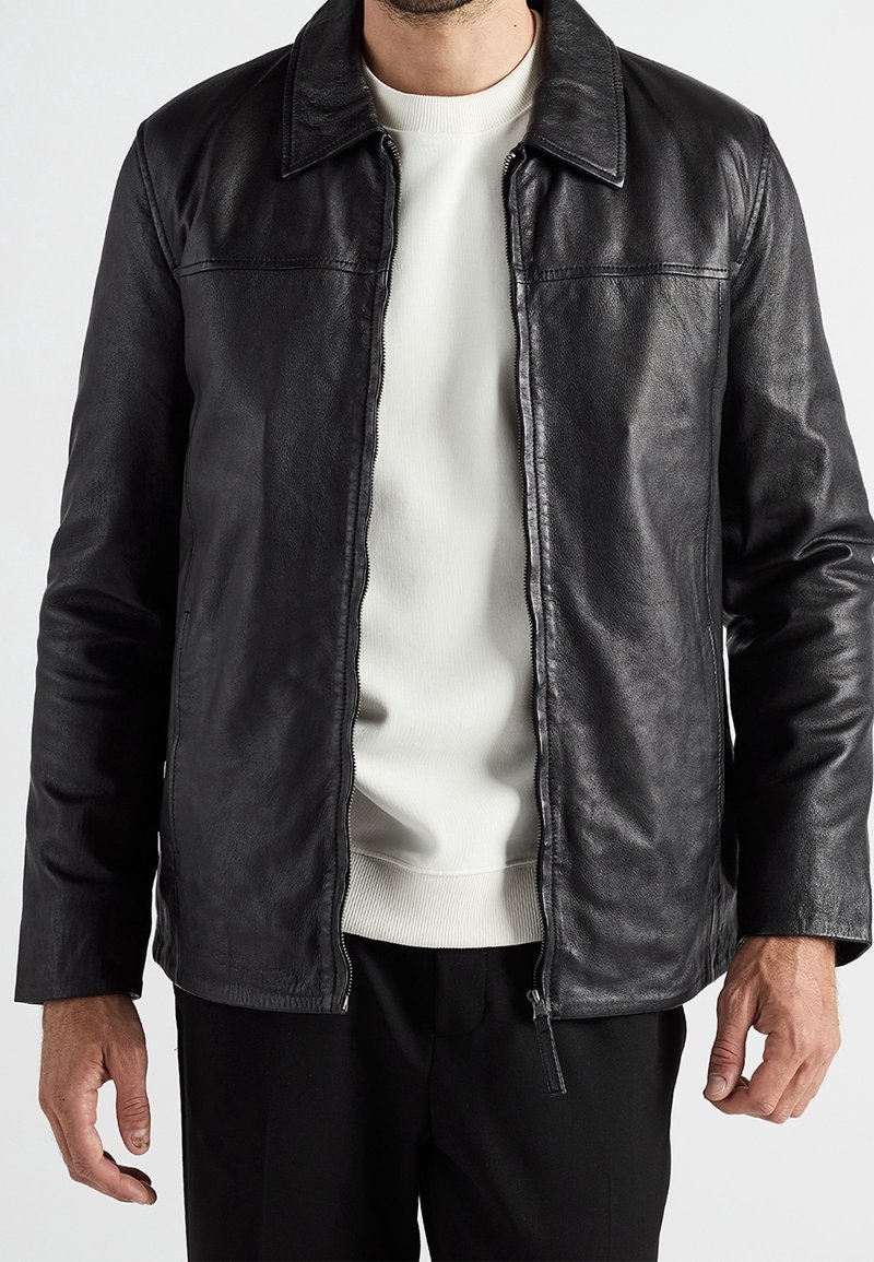 Homme portant une veste en cuir noire par-dessus un pull blanc à col rond sur un fond clair uni.