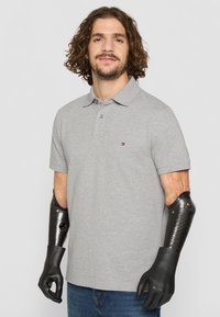 Grå polo med kort krage og tre knapper. Har et logodesign på brystet. Bæres med blå jeans og sorte protesearmer.