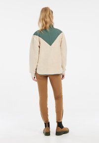 Fleecejack in roomkleur met groene kraag en een V-vormig ontwerp; gecombineerd met bruin aansluitende broek en bruine enkel laarzen. Soepele textuur.