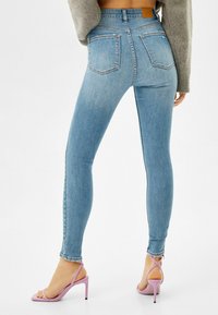 Vaqueros de mezclilla azul ajustados con una cintura alta, con dos bolsillos traseros, textura desgastada y acabado envejecido. Combinados con tacones altos de color púrpura claro.