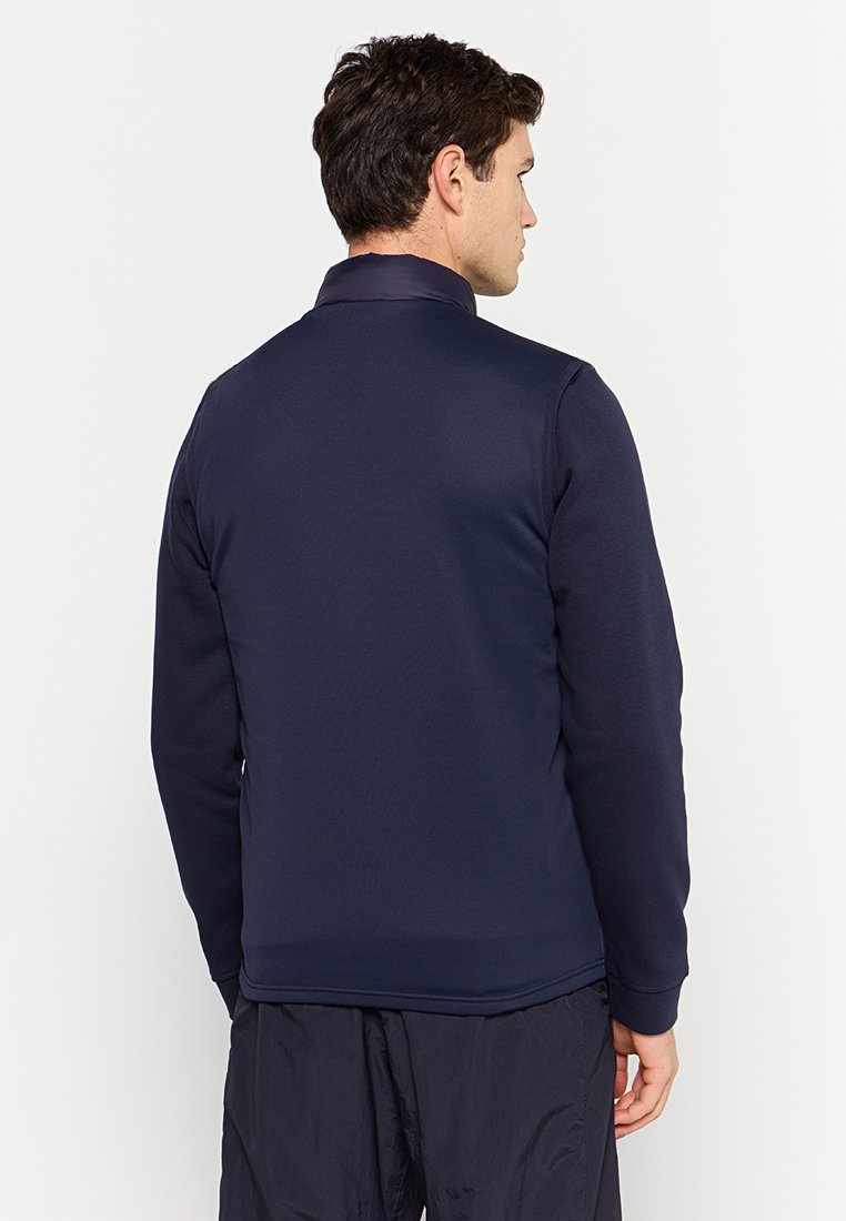 Veste en polaire bleu marine avec un col haut et une fermeture éclair. Texture lisse avec des manches longues, se prolongeant jusqu'à un ourlet arrondi à la taille.