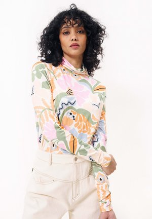 Pull à col roulé à manches longues en tissu léger, présentant un motif floral coloré avec des teintes de rose, vert et orange. Pantalons beiges unis.