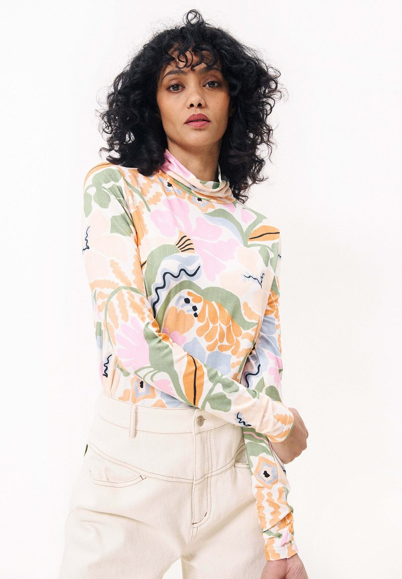 Pull à col roulé à manches longues en tissu léger, présentant un motif floral coloré avec des teintes de rose, vert et orange. Pantalons beiges unis.