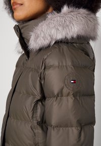 Femme portant une doudoune vert olive avec une bordure de capuche en fausse fourrure grise et un patch logo Tommy Hilfiger sur le haut du bras.