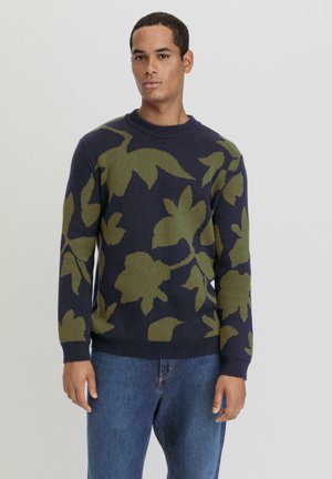 Gebreid sweater met een marineblauwe achtergrond en een groot groen bladraster, ronde hals en geribbelde manchetten. Gecombineerd met blauwe jeans.