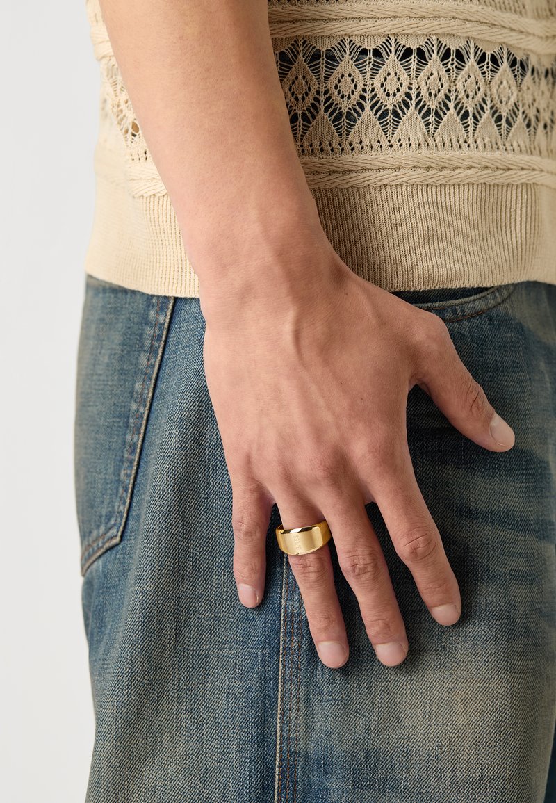 Gold-colored ring on a hand, featuring a smooth, wide band. The hand is resting on denim fabric, with visible stitching and a slightly faded texture.