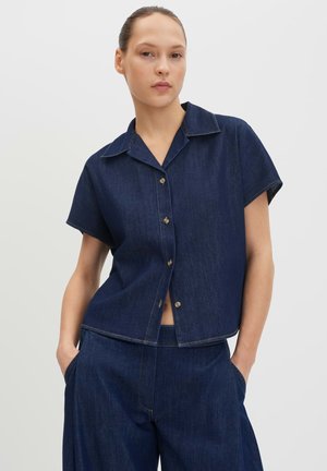Femme portant une chemise en jean bleu foncé à manches courtes avec des boutons et un pantalon en jean taille haute assorti, debout les mains dans les poches.