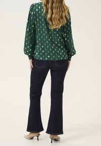 Blusa verde con motivi a foglia dorata, maniche ampie e polsini arricciati, abbinata a jeans svasati scuri e mule beige con tacco.