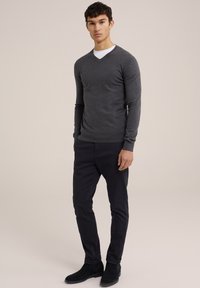 Grauer V-Ausschnitt-Pullover mit langen Ärmeln, taillierte Passform, über einem weißen Hemd getragen, kombiniert mit dunklen Hosen und schwarzen Stiefeletten.