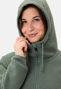 Giacca a vento verde in fleece con chiusura a zip, superficie testurizzata, cordoncini regolabili e rifiniture in contrasto attorno ai polsi e alla zip.