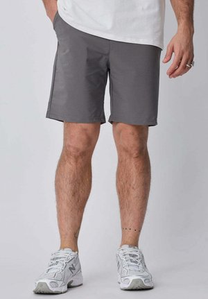 Homme portant un short gris, des baskets blanches et une chemise blanche, debout avec une main dans la poche devant un fond uni.