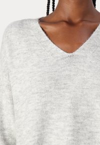 Jasnoszary sweter wykonany z miękkiego materiału z głębokim dekoltem w kształcie litery V. Ukazuje teksturę i niewielkie różnice w kolorze na tkaninie.