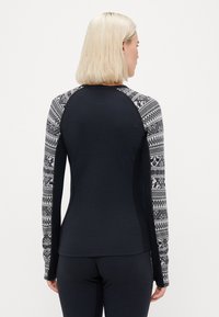 Žena s krátkymi blond vlasmi, oblečená v čiernom topu s dlhými rukávmi, ktorého rukávy sú čiernobielo vzorované, stojí otočená chrbtom k bielemu pozadiu.
