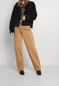 Svart sammetsjacka med huva, en mönstrad skjorta under och ljusbruna vida jeans. Svarta ankelboots fulländar outfitten.